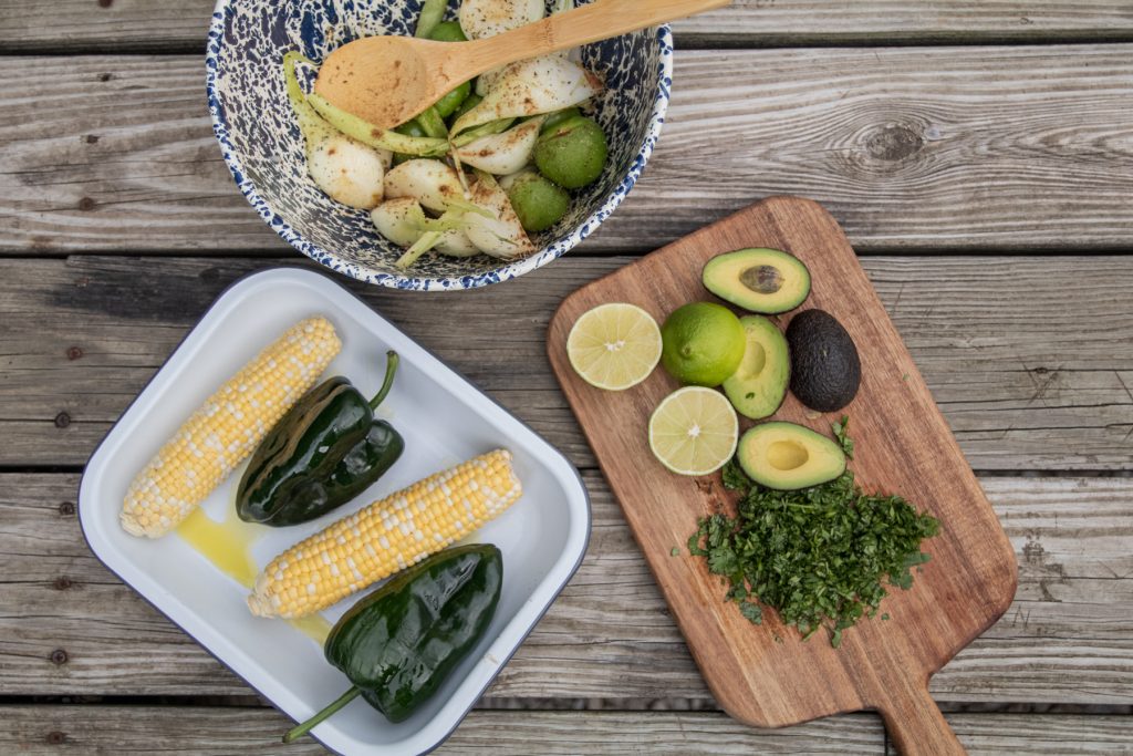 A bowl full of onions and tomatillos, a platter full of poblano peppers and ears of corn, and a cutting board with cilantro and avocado on a picnic table.