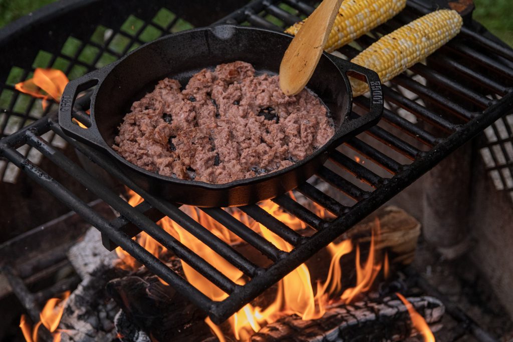 Ears of corn grilling on top of the campfire next to a Lodge cast iron pan cooking Beyond Meat beef crumbles.