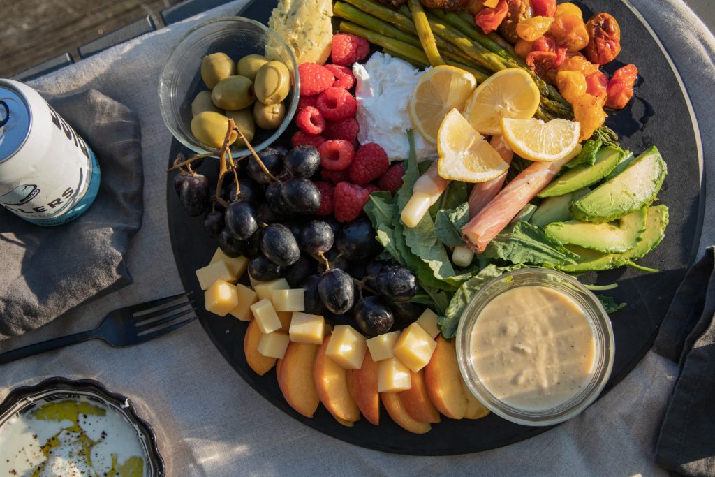 A closeup of a platter full of peaches, cheese, grapes, olives, raspberries, lemons, tomatoes, asparagus, and avocado with a side of hummus.
