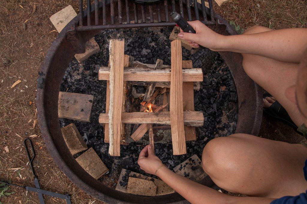 A woman lights the tinder and kindling of a log cabin structure made from campfire wood.