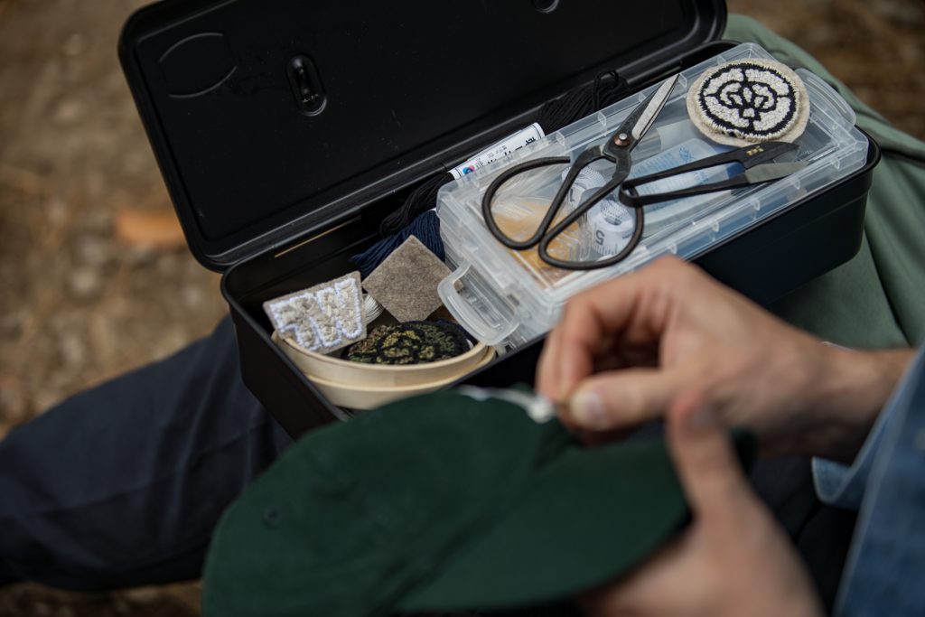 A man embroiders his hat with his case full of threads and patches sits on his lap.