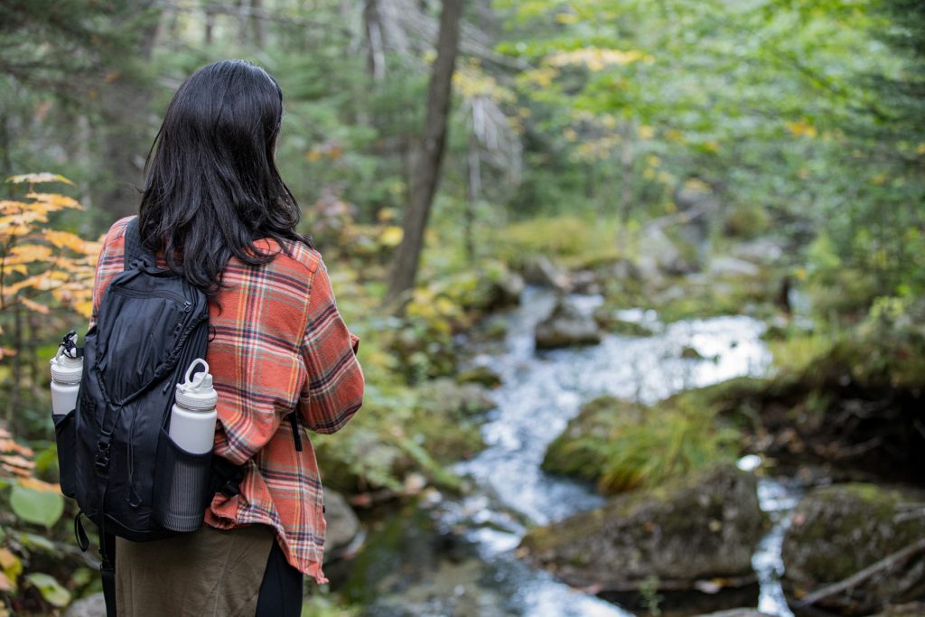 A woman hiking in Maine wearing LL Bean's classic plaid flannel, wearing a backpack with two white Takeya water bottles, and overlooking a creek.
