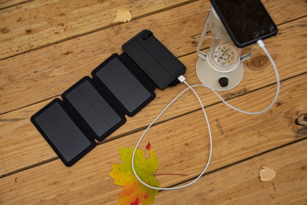 A portable solar panel phone charger on top of a wooden picnic table. A phone is connected to the charger.