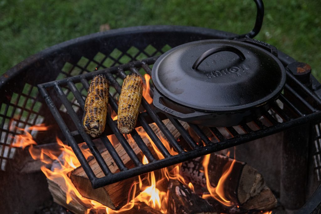 A Lodge Cast Iron double-handle pan with a lid on top of a campfire grate next to roasted corn on the cob.