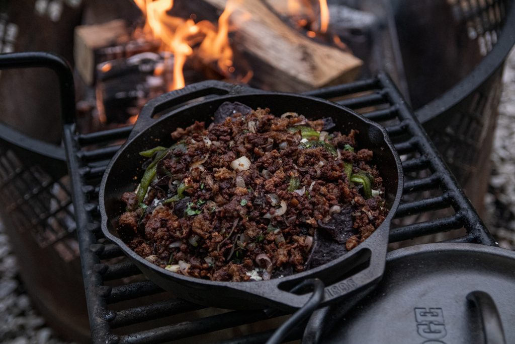 A Lodge Cast Iron double-handle Pan with nachos and ground beef with a campfire in the background.