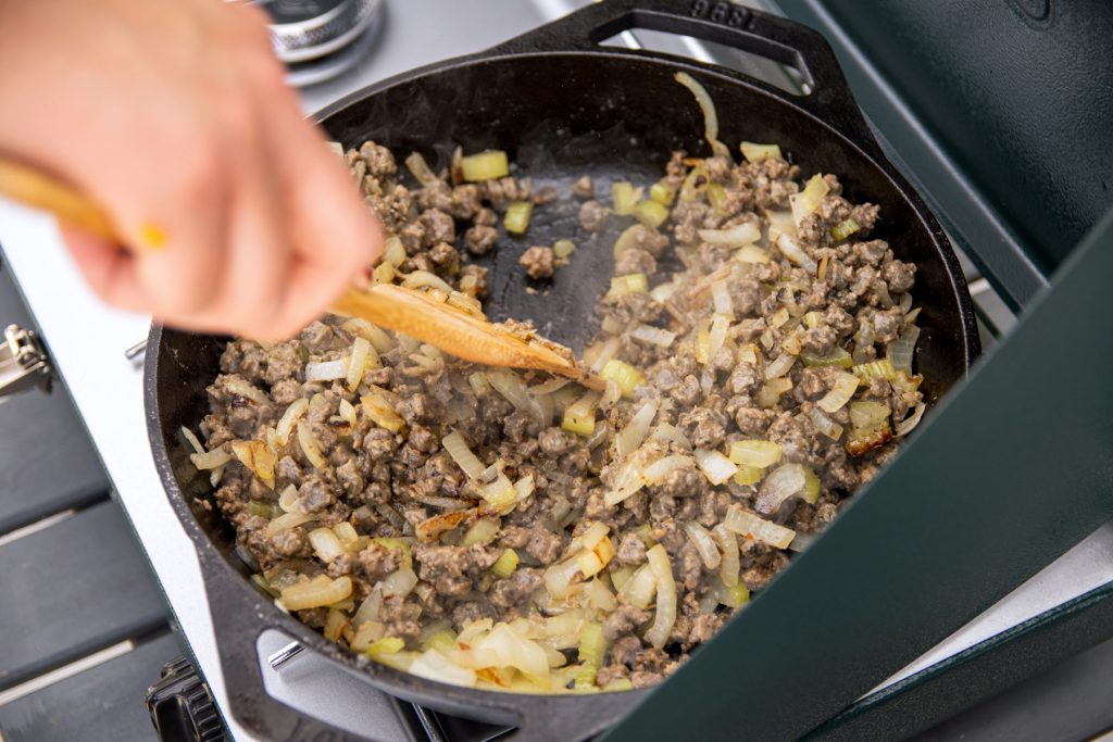 A person stirs a combination of cut onions, sausage, and sausage crumbles within a Lodge cast iron pan on a Coleman camp stove. 