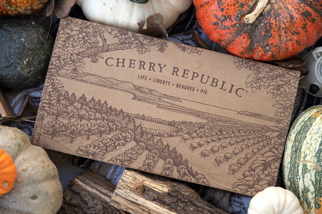 A Cherry Republic box with an illustration of cherry trees overlooking Lake Michigan is surrounded by decorative squash and pumpkins.