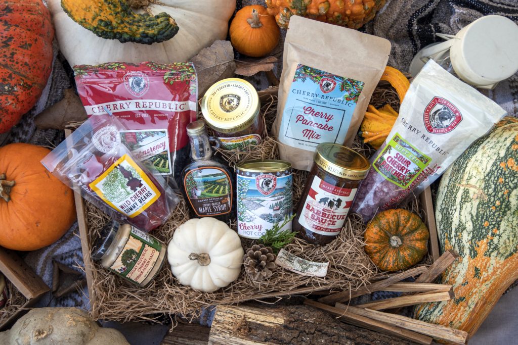 A box full of Cherry Republic merchandise is surrounded by decorative squash and pumpkins. Inside the box includes items such as Cherry hot cocoa, Cherry candy, Cherry pancake mix, and more.