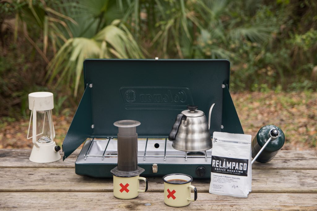 Coleman Portable propane gas classic two-burner stove on top of a picnic table with coffee mugs, an aero press, water kettle, and bag of coffee surrounding it.