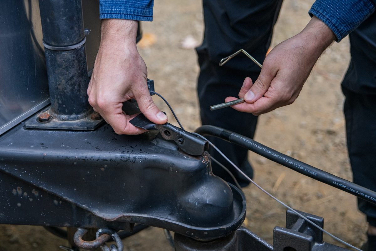 A person is securing the breakaway switch between the tow vehicle and trailer. A breakaway switch is a feature that activates the trailer's breaks if it disconnects from the tow vehicle in the case of an emergency.