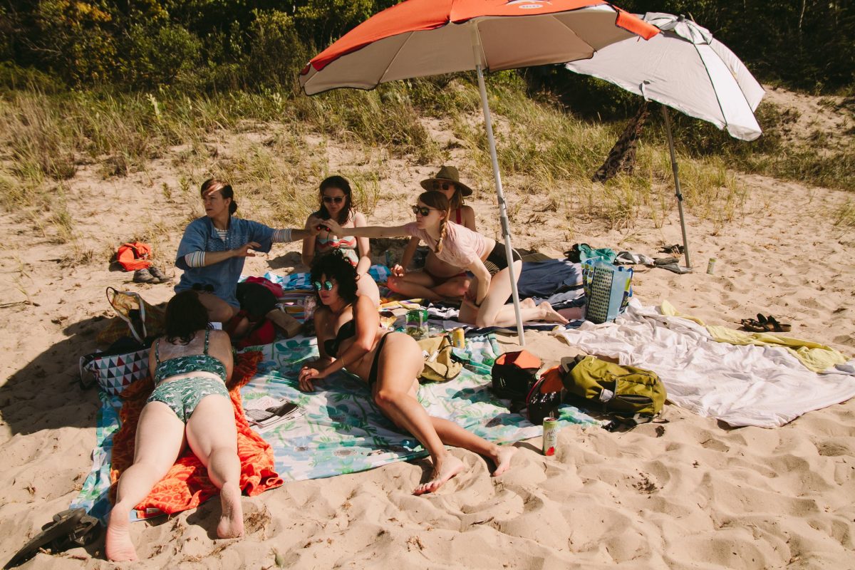 A group of six women lounging in the sun on beach towels, with two umbrellas propped open for shade.