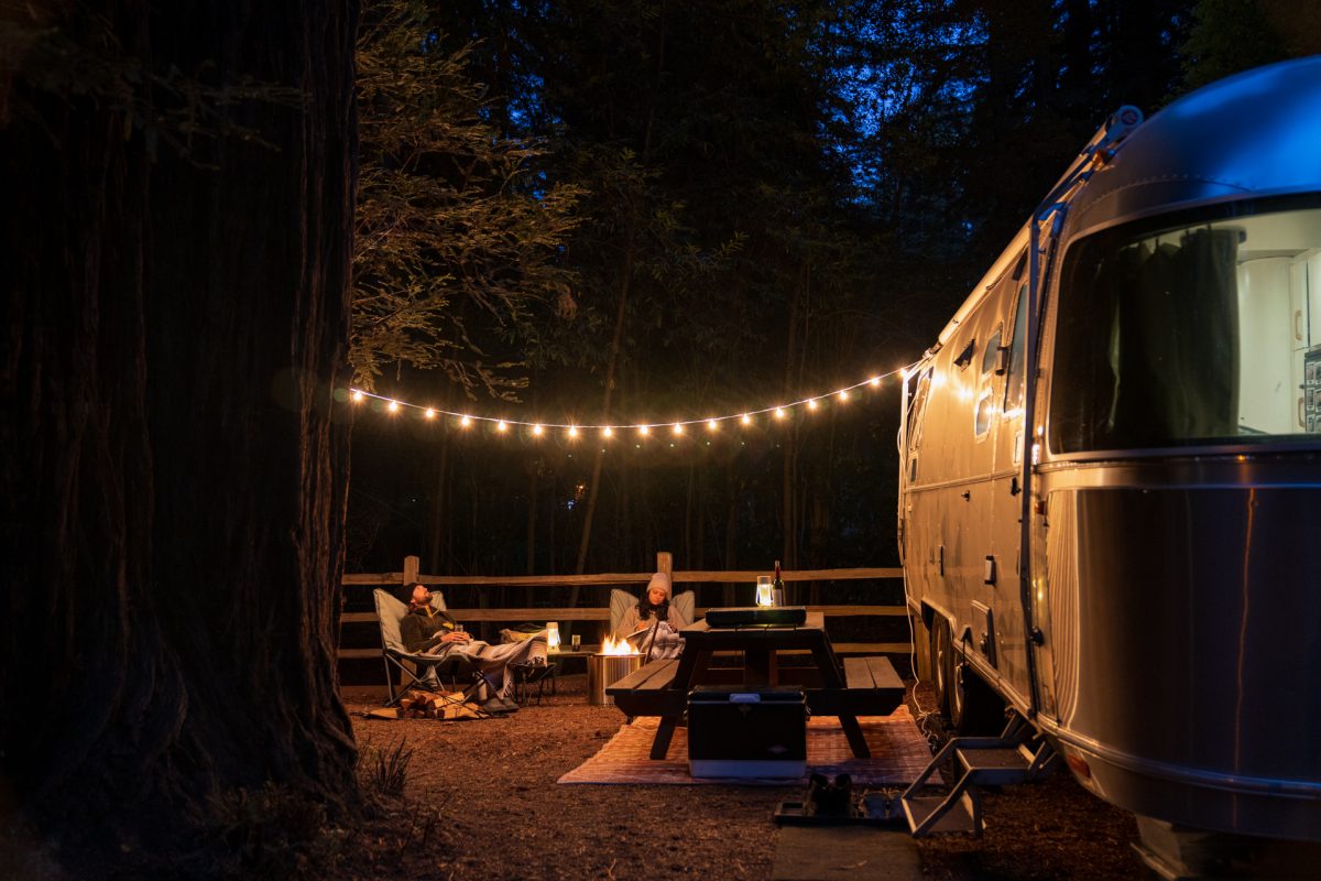A couple sits at their campsite at the Santa Cruz Redwoods RV Resort in Felton, California. It's nighttime and their string lights sit above them and light up the redwoods around.