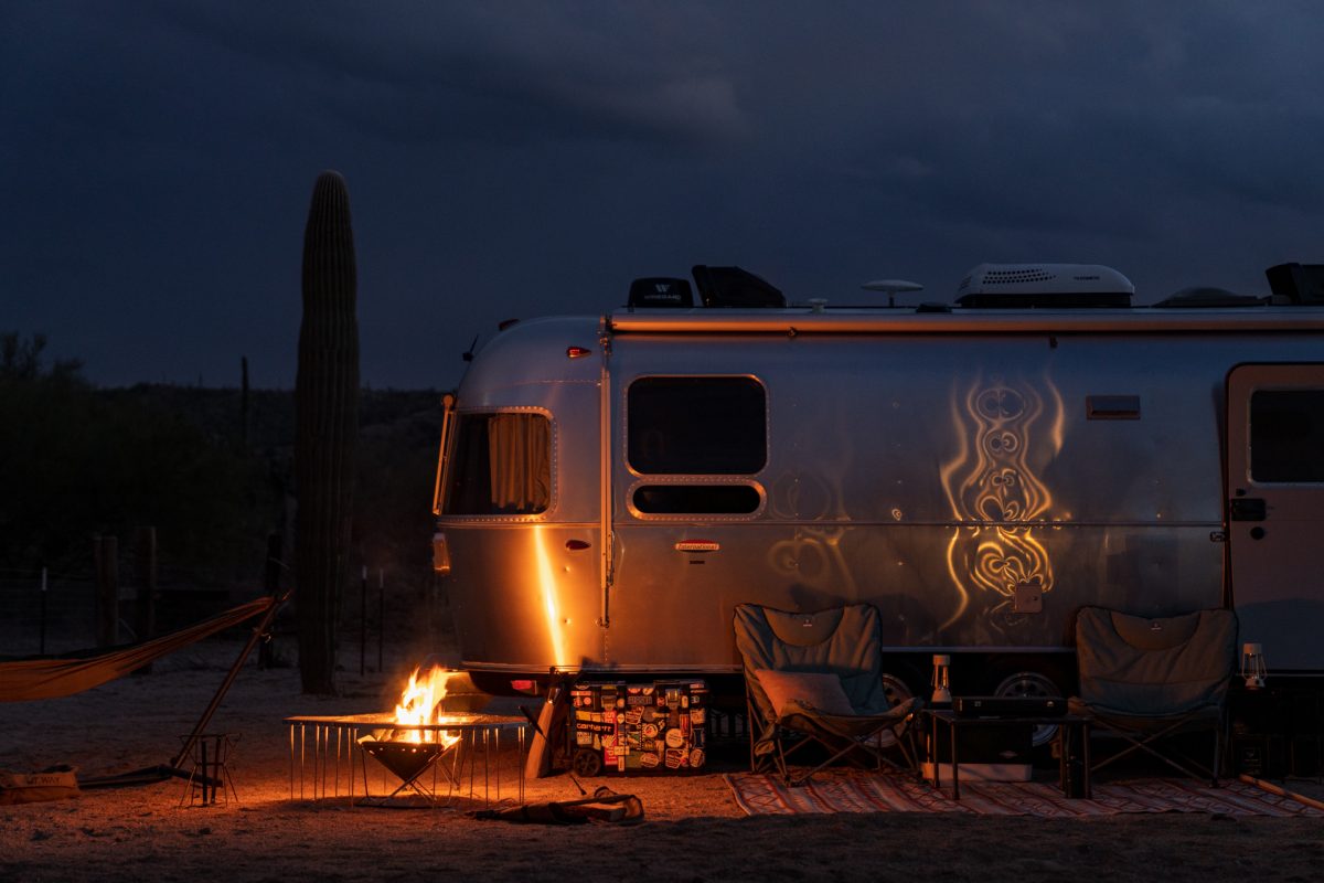 An Airstream RV and travel trailer sits at its sandy campsite with a campfire going in Ride Out Ranch Campground in Florence, Arizona.