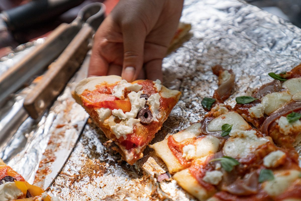 A person picks up a slice of campfire pizza that has goat cheese, olive, ans tomato sauce. 