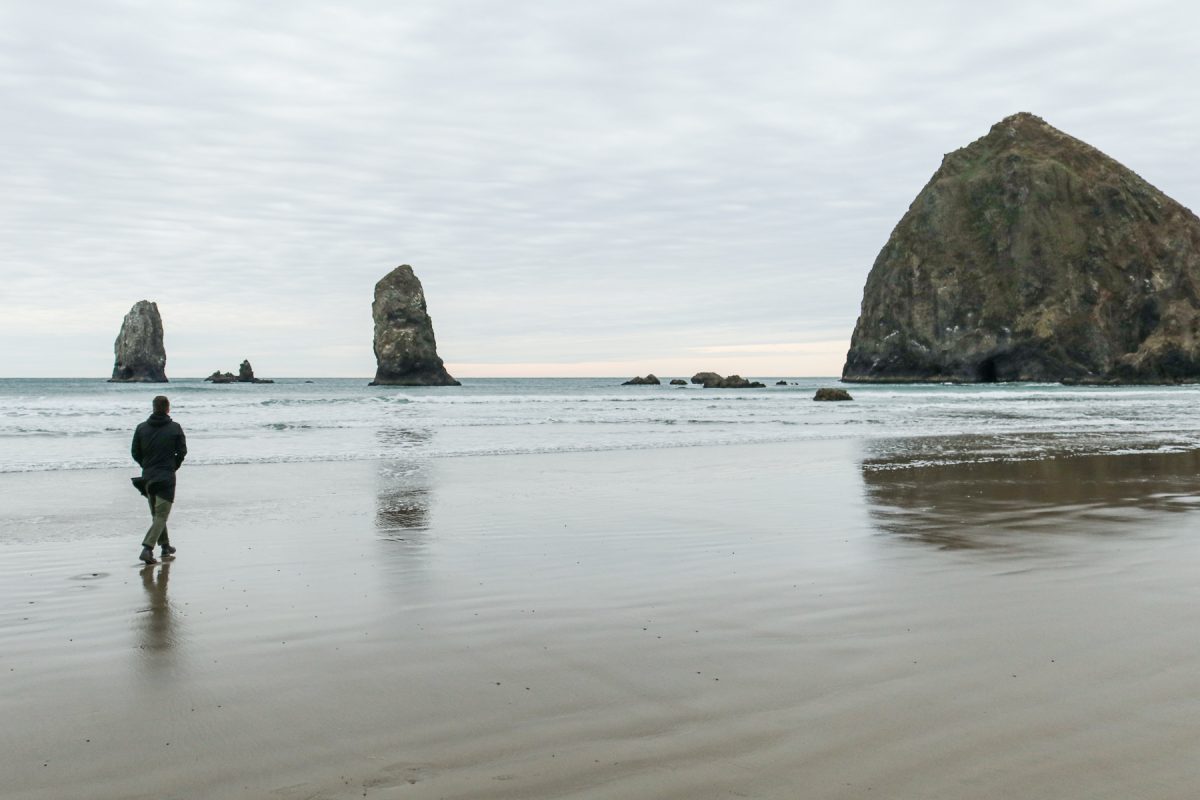 A person walks Cannon Beach with the Needles and the Haystack Rock in the backdrop.