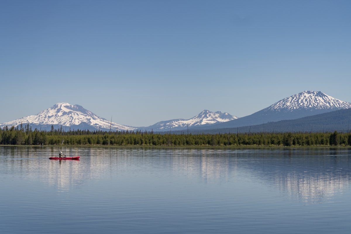 A person in a red kayak glides across one of Oregon's Cascade Lakes with the Three Sister Mountains in the background. The Cascade Lakes is a great destination for those on an Oregon road trip. 