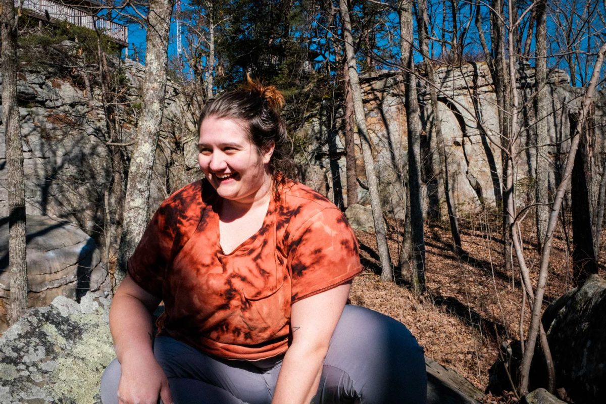 A person smiles while seated in a wooded area. 