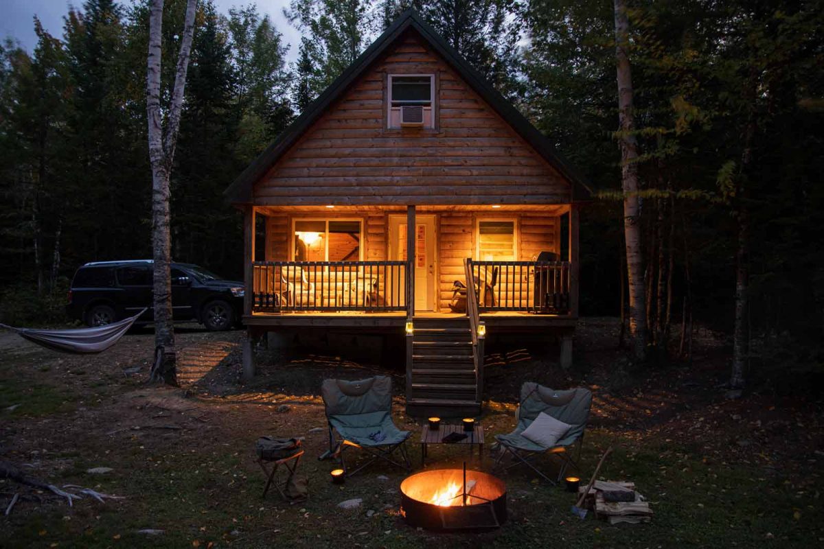 An a-frame cabin at night with a fire pit and two chairs in front of the porch. 