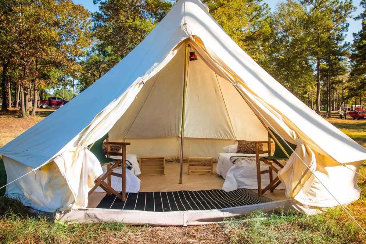 A cream-colored glamping tent with two twin beds and a rug inside with trees in the background. 