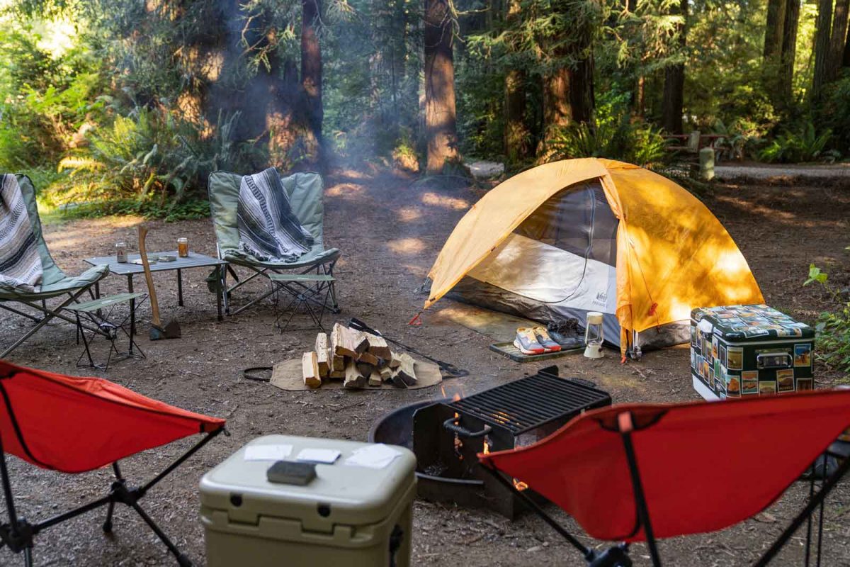 A frontcountry tent camping 101 for beginners set up that includes a tent, cooler, folding chairs, table, and campfire. 