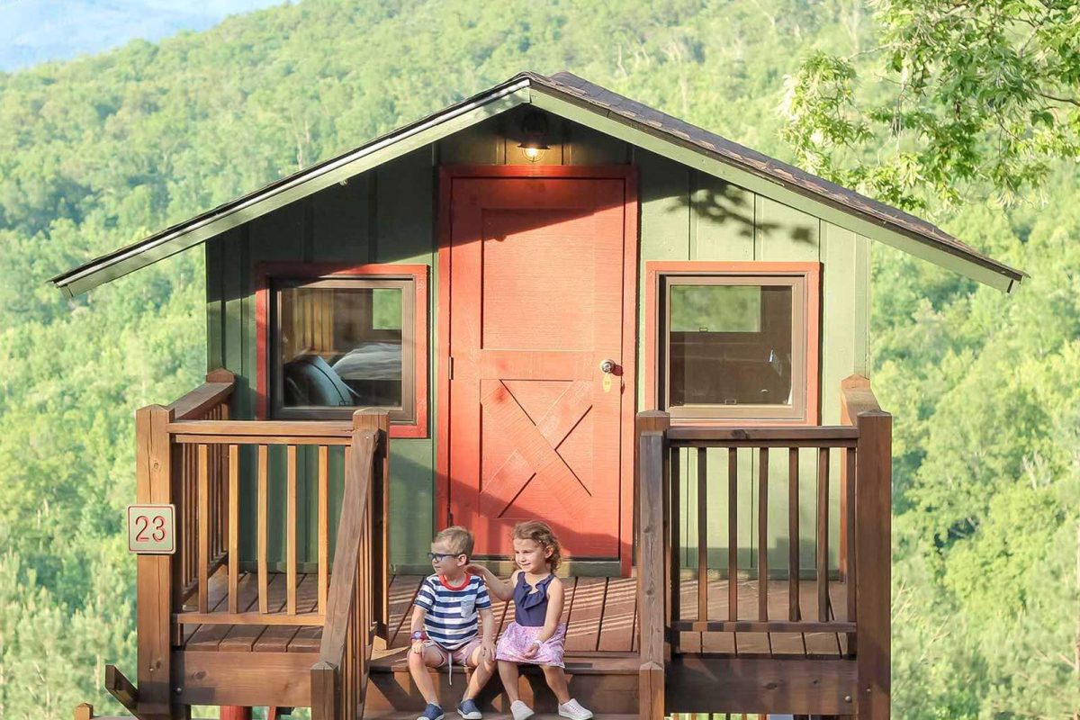 Two young children sit on the front porch of a colorful cabin at Yogi Bear’s Jellystone Park™ Camp Resort: Golden Valley. 