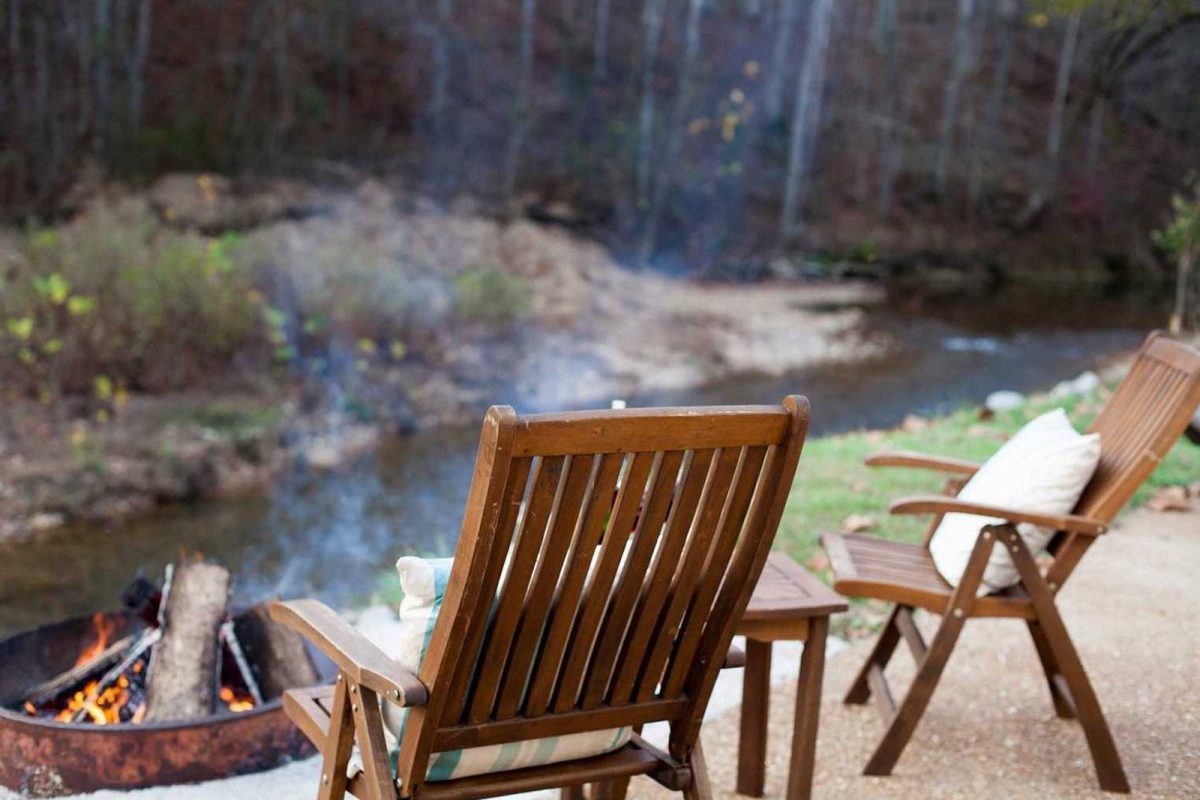 Two wooden lounge chairs face a fireplace and riverbank at Piney River RV Resort, a campground off the beaten path. 