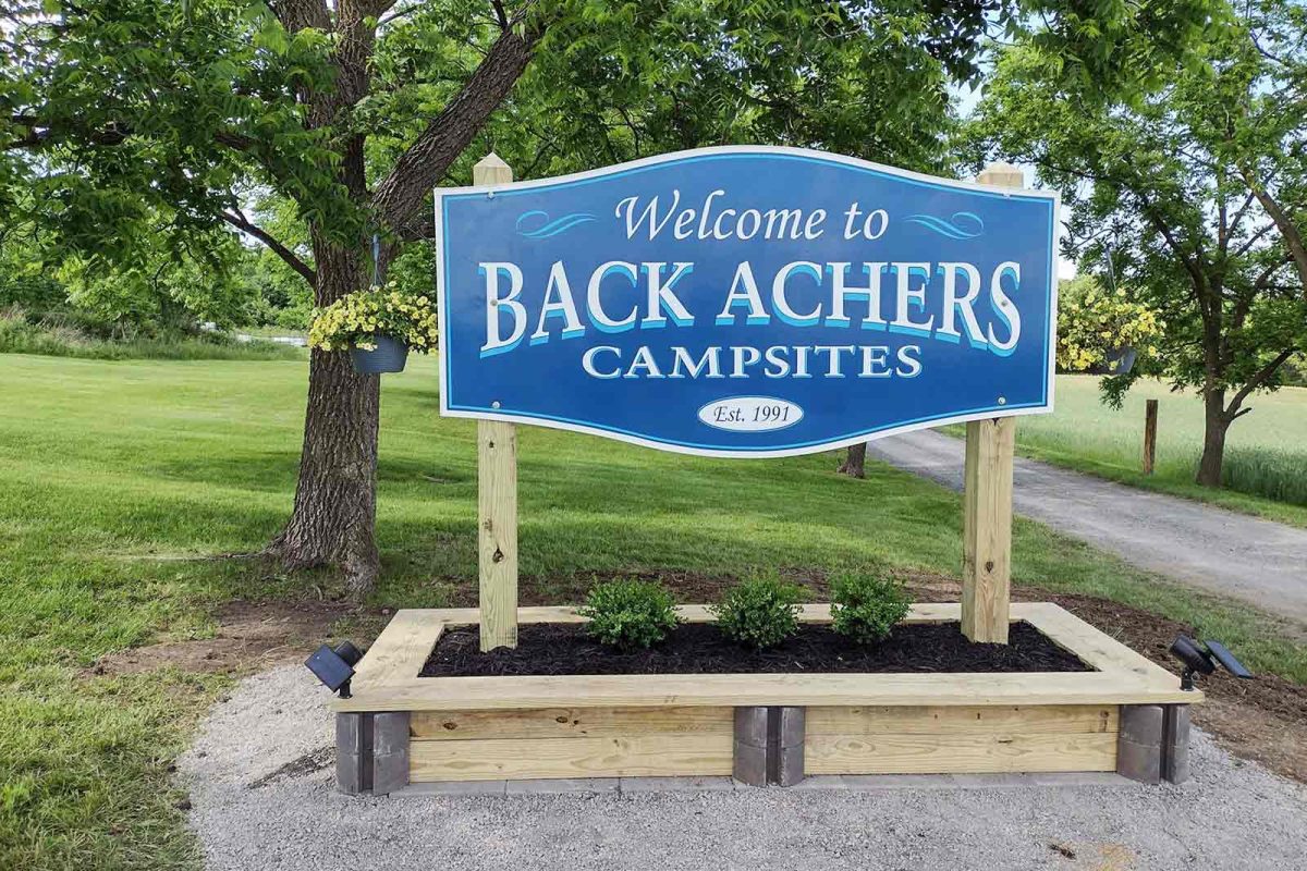 The blue welcome sign at Back Achers Campsites reads, "Welcome to Back Achers Campsites Est. 1991." 