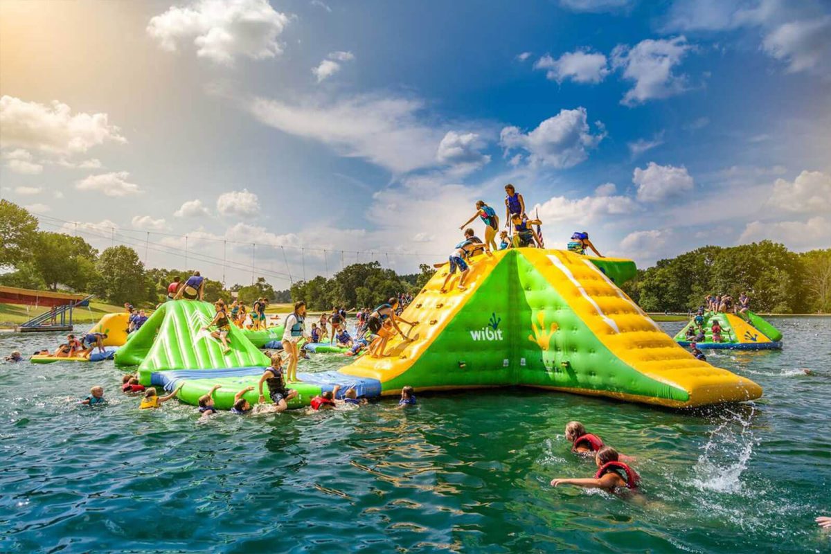 Campers climb all over a yellow and green inflatable water slide in the water at Yogi Bear's Jellystone Park™ Camp-Resort: Clay's Resort, one of the campgrounds with water parks on Campspot. 