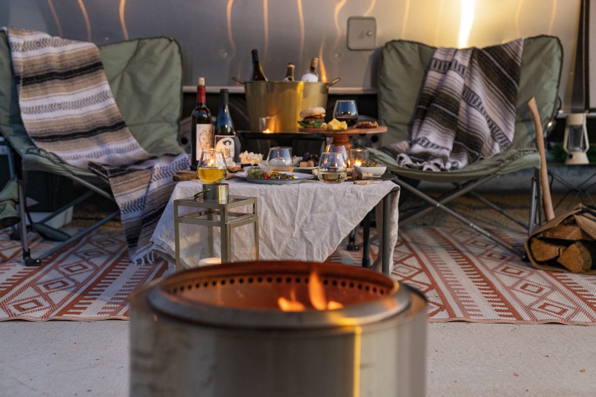 Two camp chairs with blankets sit on either side of a table filled with wine and snacks. A fire burns in the forefront. 