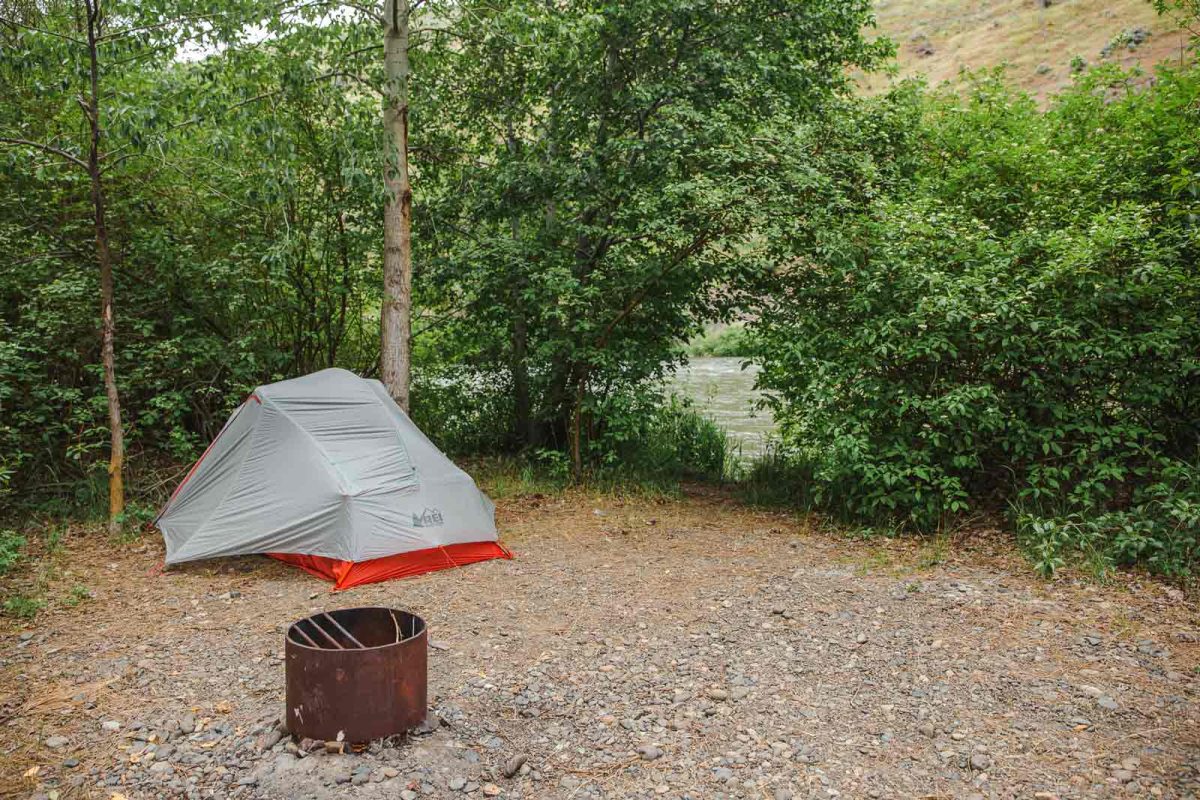 A grey and white tent next to a fire pit and riverside view. 