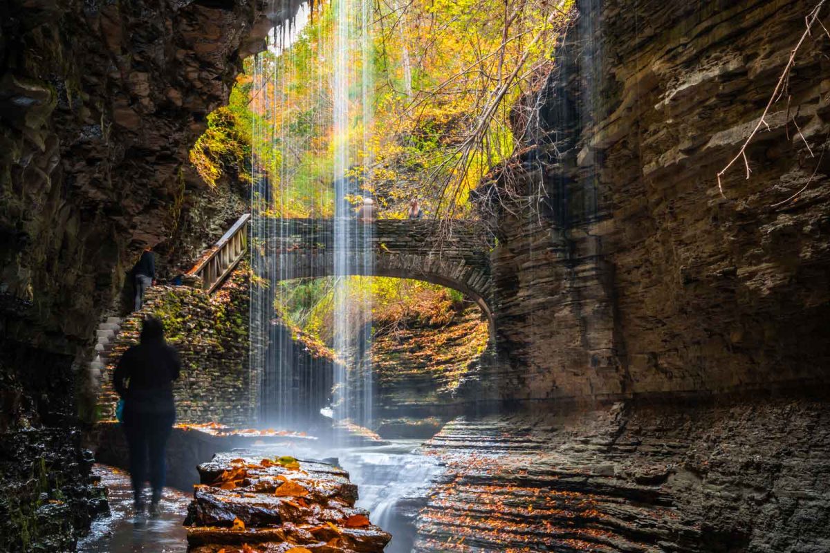 A hiker stands on a trail beneath a waterfall and bridge in the distance with fall foliage on the trees 