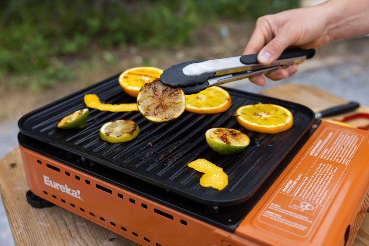 Hand holds tongs that grip charred slices of citrus on a camp stove grill. 