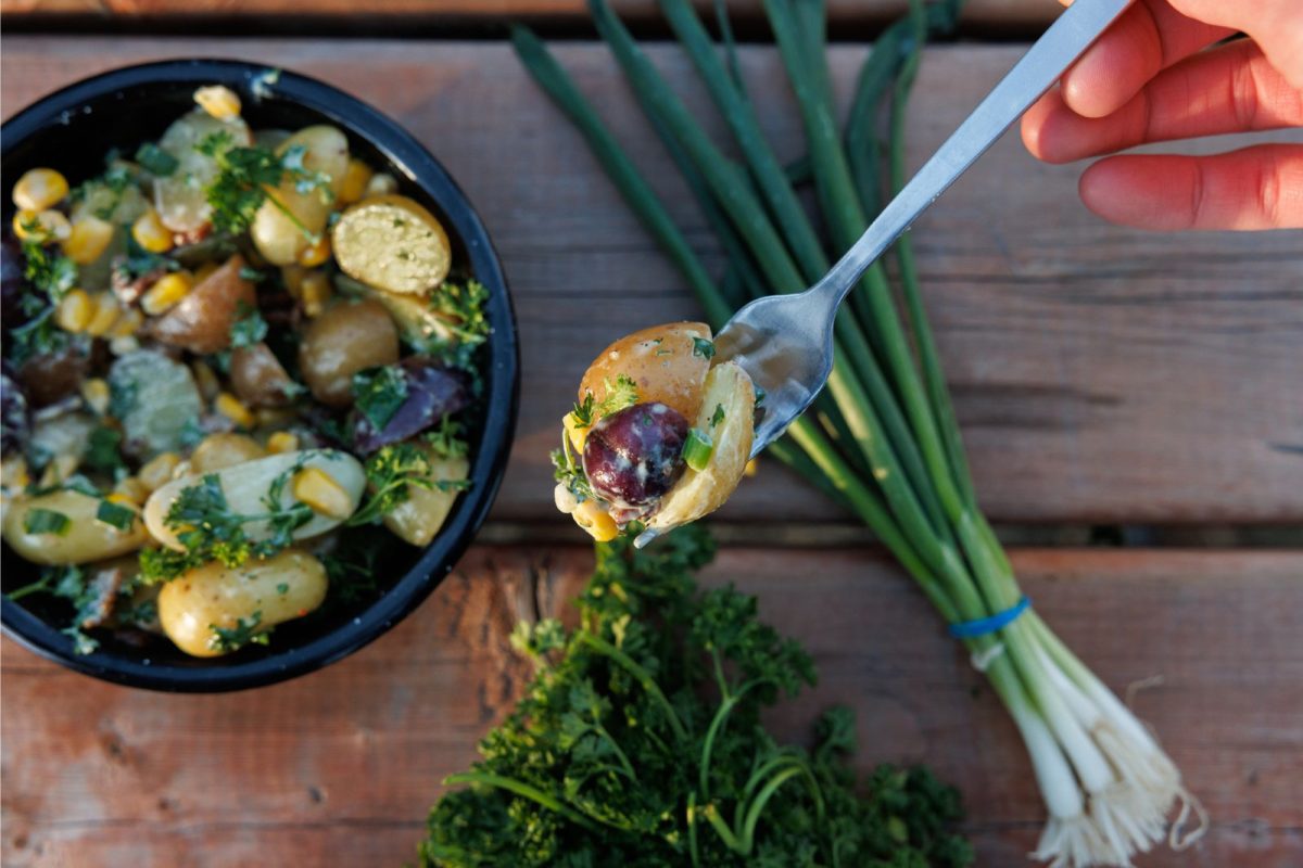 person holding up a fork of baby potato with olive, and bowl of potato salad, green onion, parsley in background