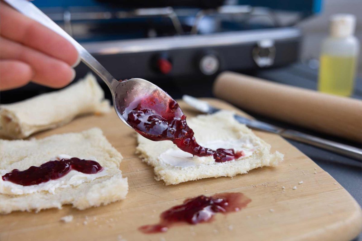 A spoon places jam on a thin piece of bread. 