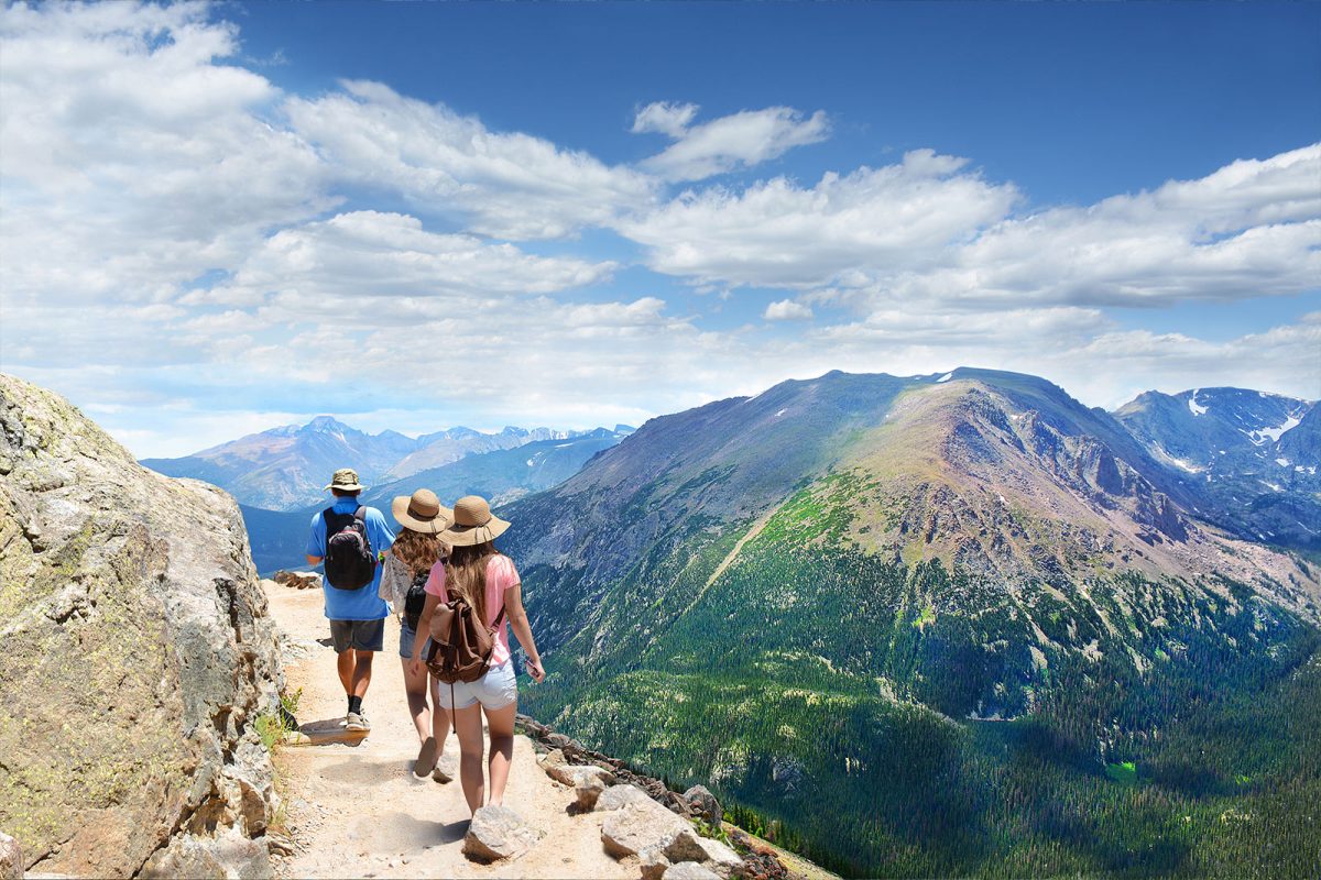 Three hikers walk a mountain-side trail with mountain views in the distance and blue skies above at Rocky Mountain National Park. 