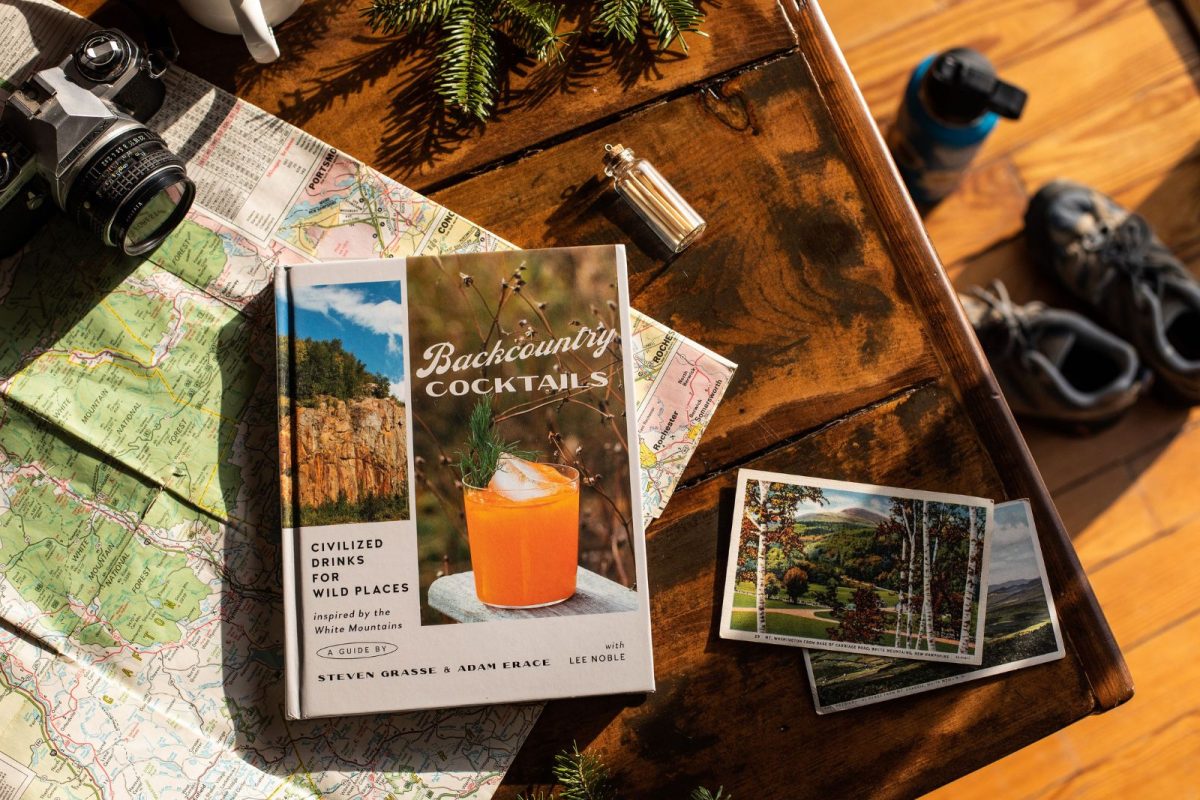 An overhead shot of the cover of the Backcountry Cocktails book. The book sits on a map on a wooden table and is surrounded by a camera, post cards, matches, and greenery. 
