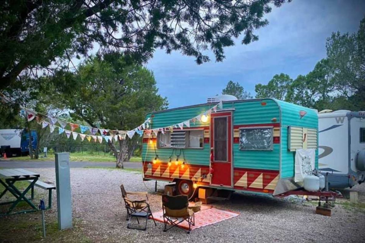 A campsite outside of a teal and red antique camper with colorful, decorative flags and twinkle lights. 