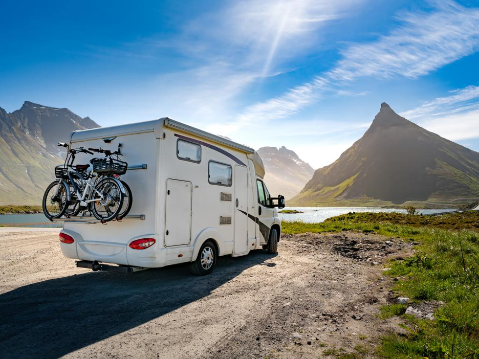 An RV with bikes on the back parked on the water's edge with mountains in the distance. 
