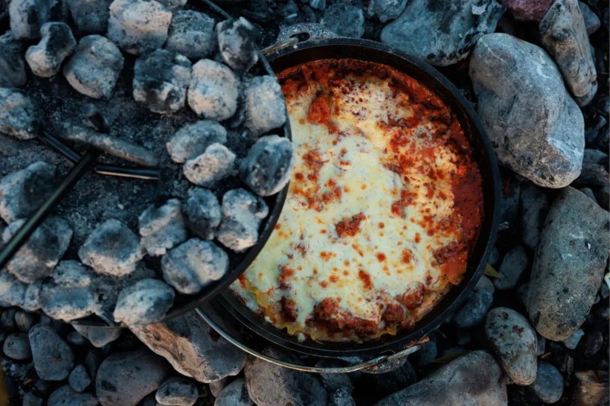 the lid being lifted off of a Dutch oven full of lasagna over the campfire